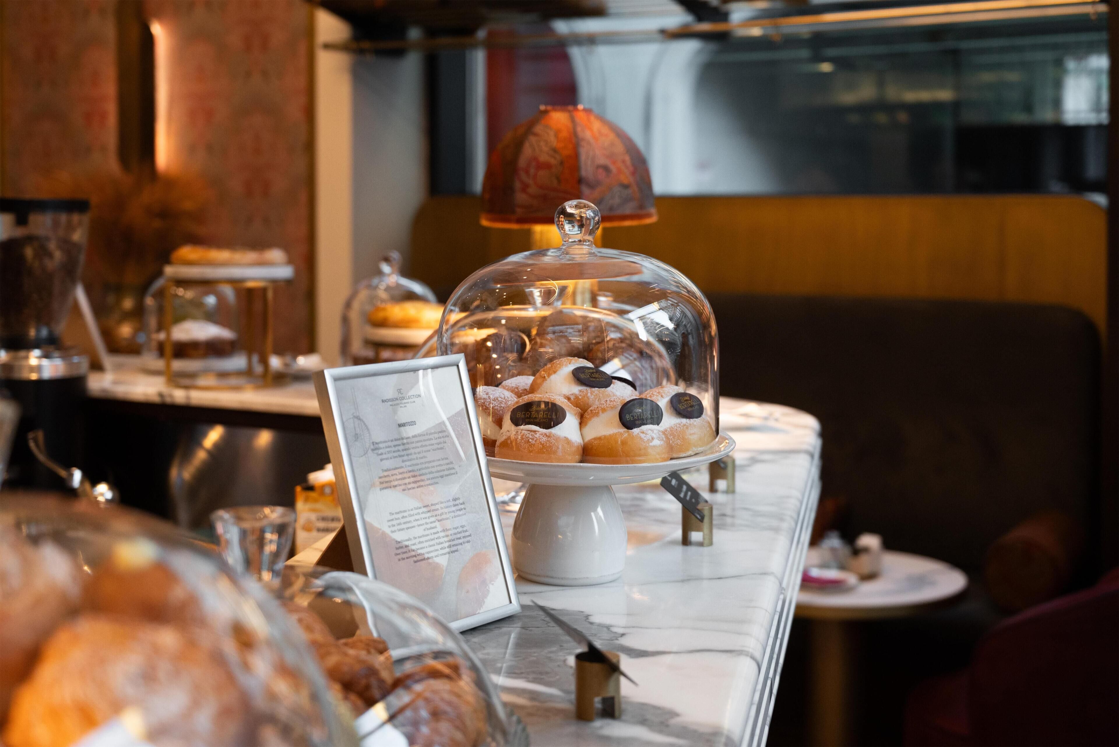 Una vetrina di pasticceria con dolci sotto una cupola di vetro, un espositore per il menu e una macchina per il caffè sullo sfondo.