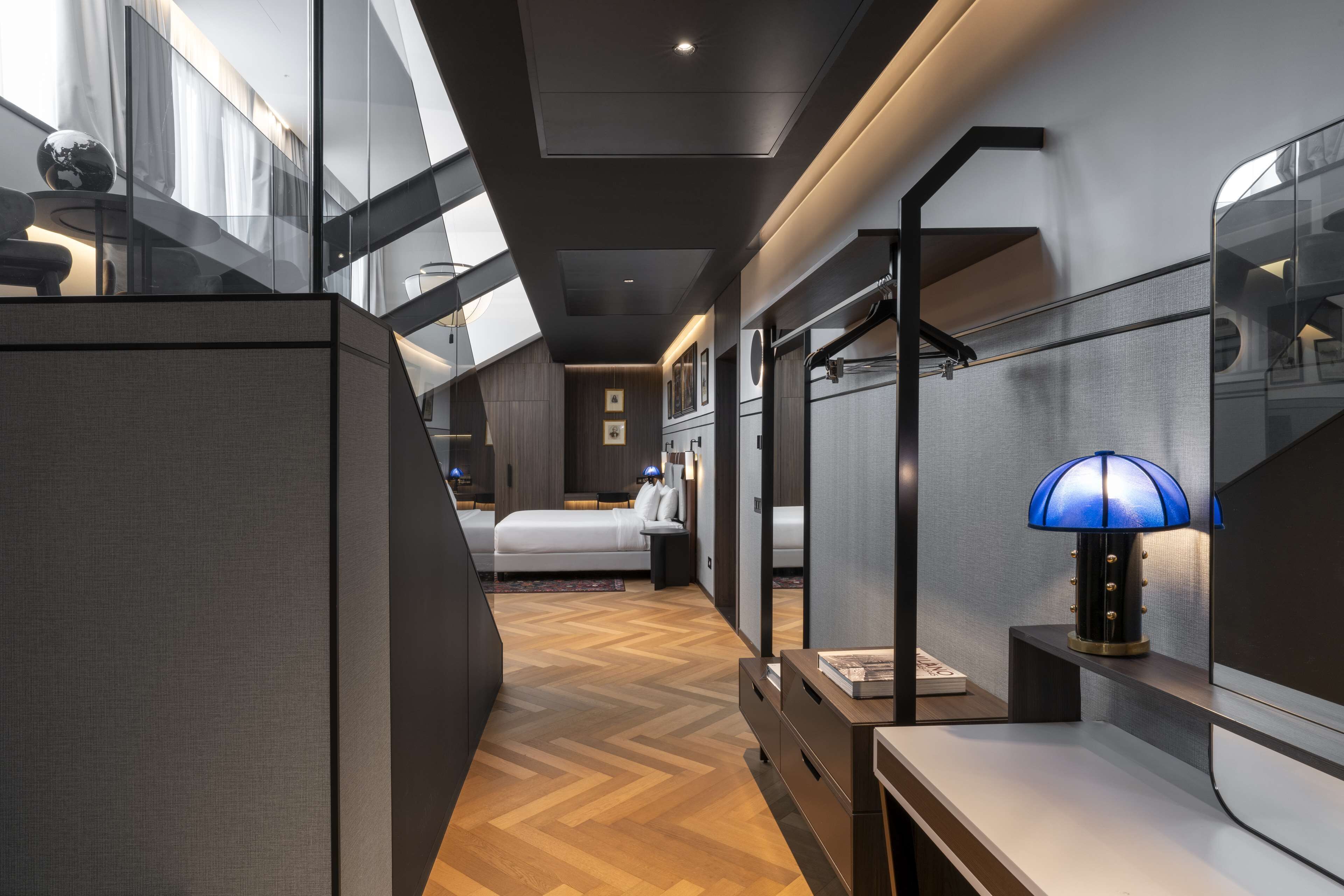 A modern hotel room with a large bed, a desk, a lamp, and a view through a glass wall.