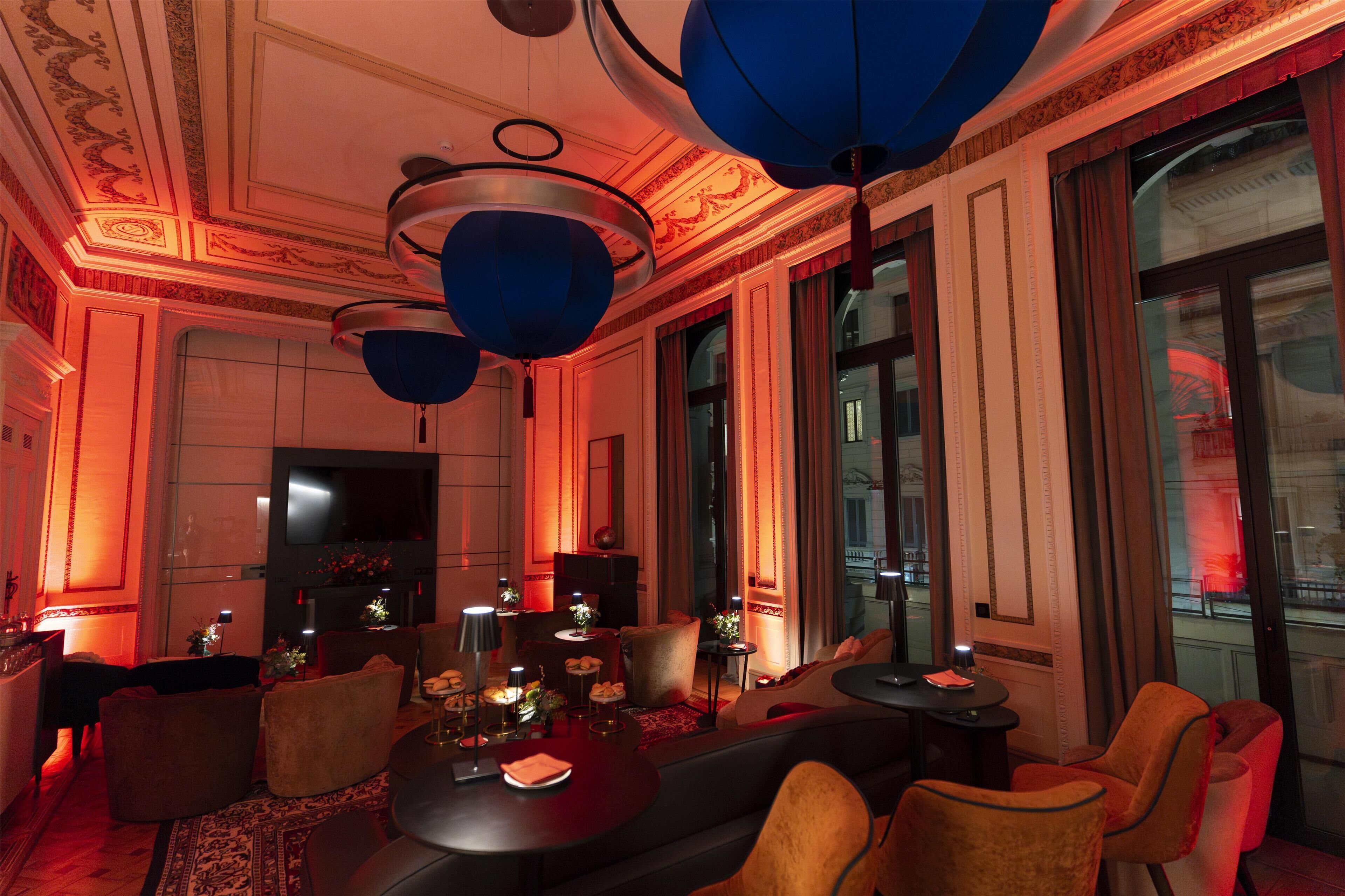 A luxurious room with a large sofa, a round table, and a television. There are multiple chairs and a small table with decorative items. The room has ornate ceiling details and large windows.