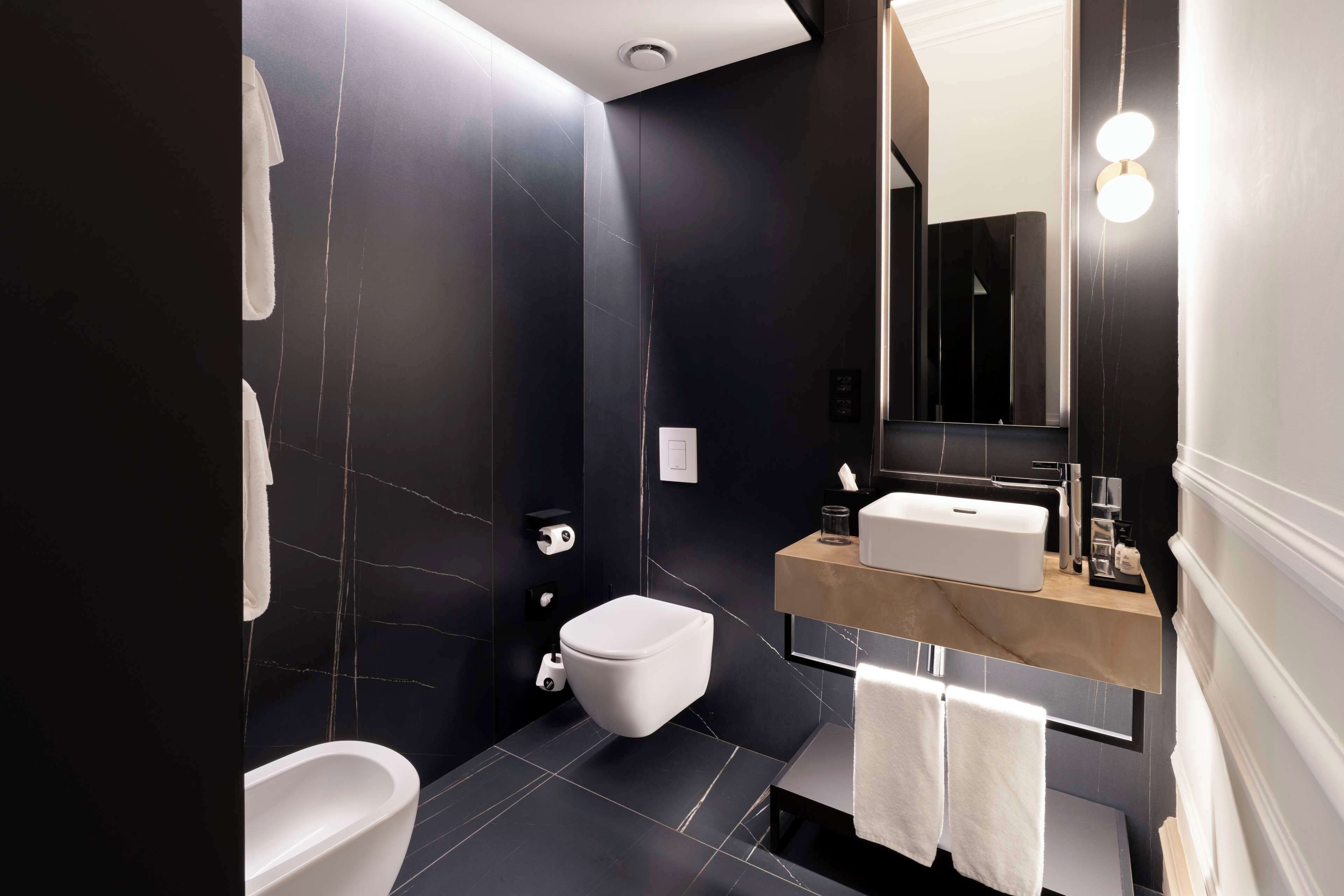 Une salle de bain moderne avec un mur en marbre noir, des toilettes blanches, une baignoire et un meuble-lavabo avec un miroir et un évier.