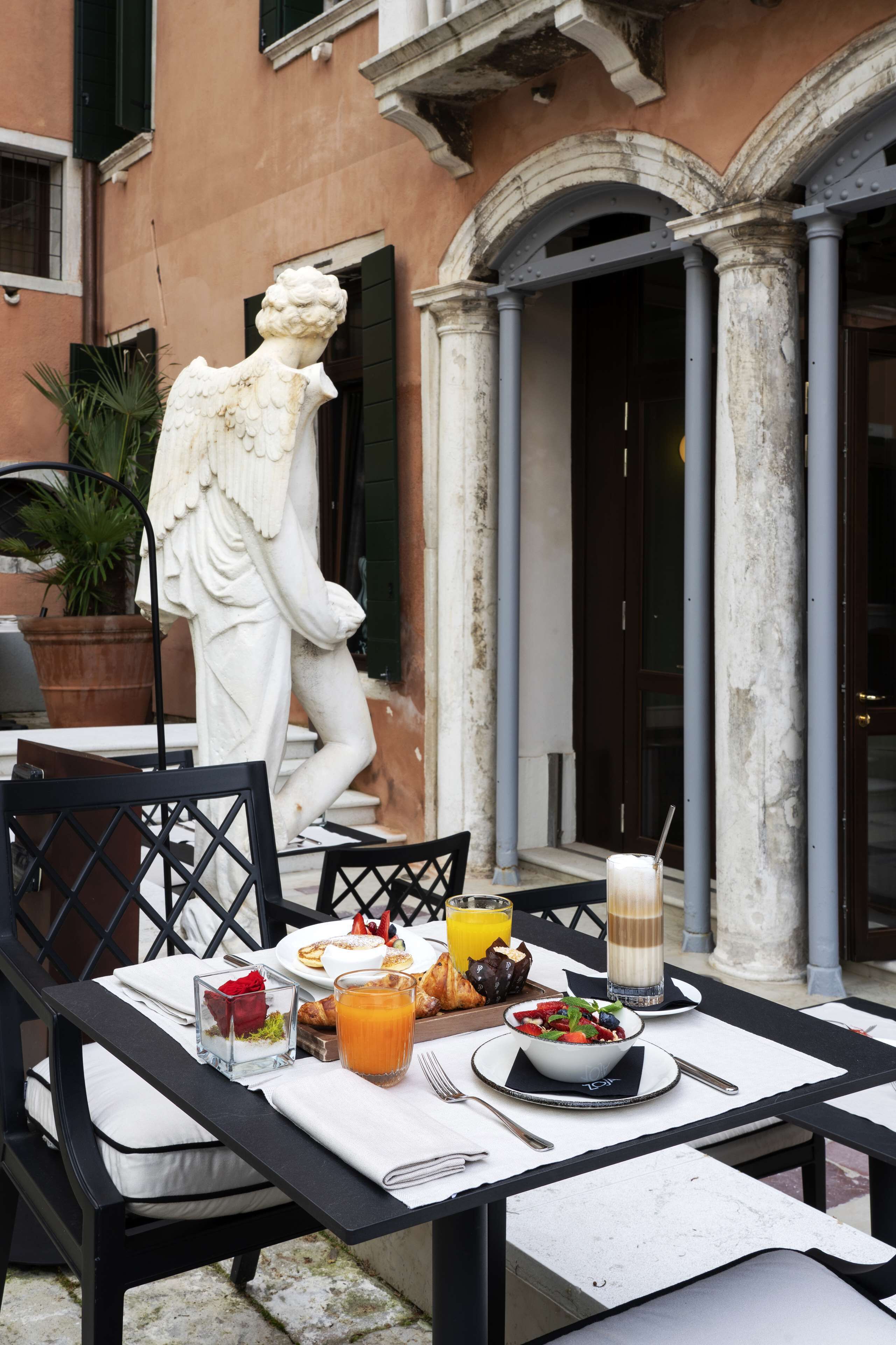 A table set outdoors with a statue of a figure, a potted plant, and a meal including a bowl of salad, a glass of orange juice, and a coffee drink.