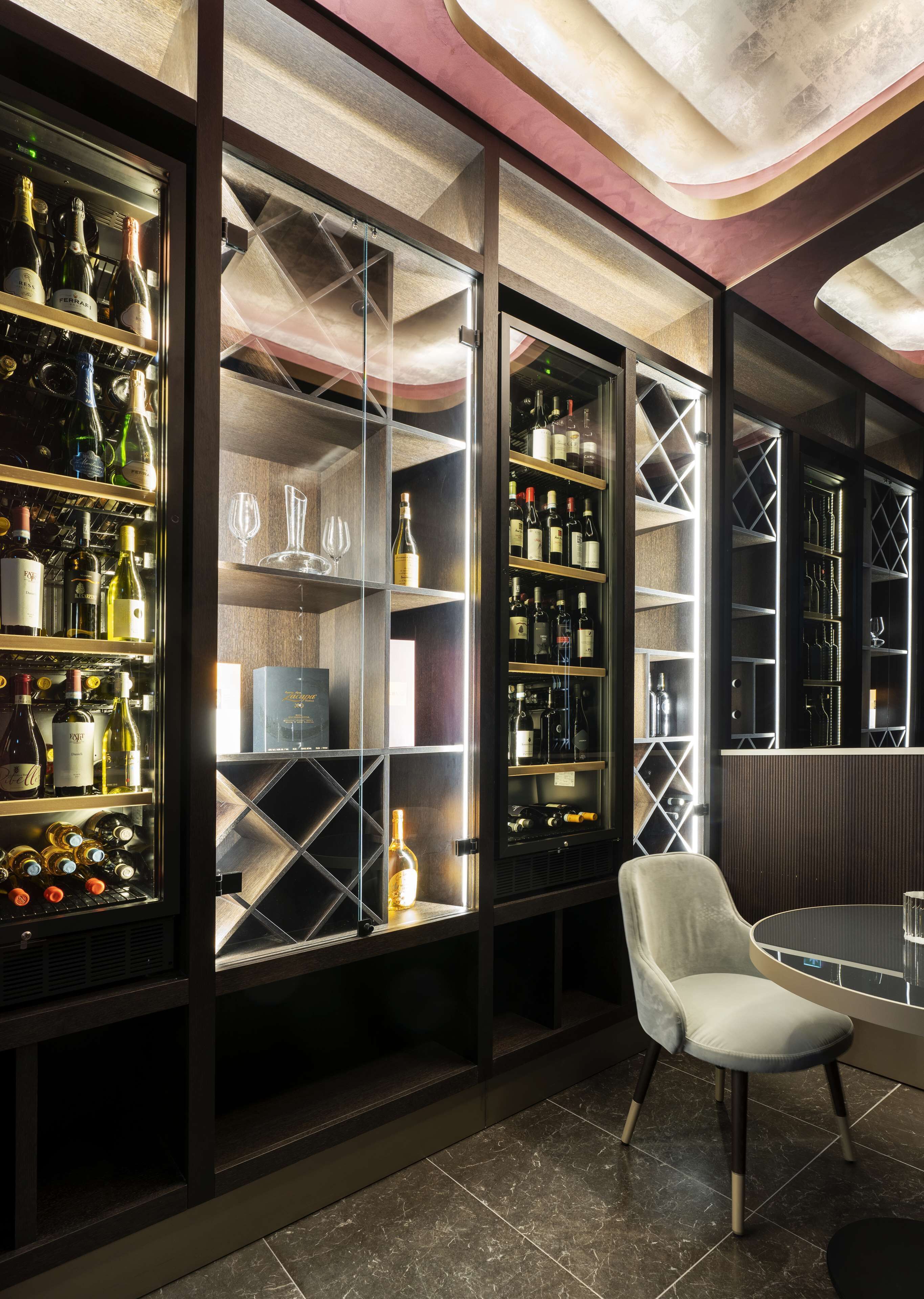 Un'area bar moderna con scaffali per vino e liquori, una zona salotto con una sedia bianca e un tavolo in vetro.