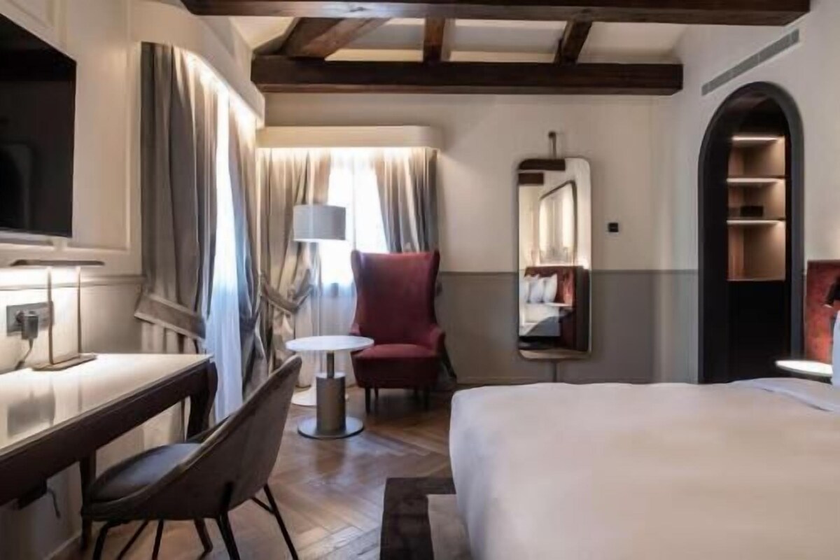 A hotel room with a bed, a desk, a chair, a small table, a mirror, and a bookshelf.
