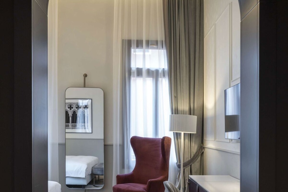 A modern hotel room with a herringbone wood floor, a red armchair, a white bed, a desk, and a large mirror.