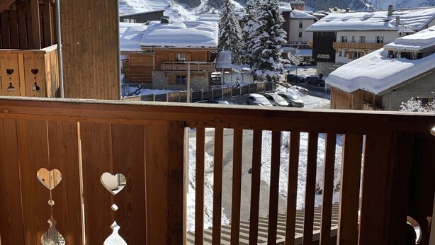 Un balcón de madera con decoraciones con forma de corazón, con vistas a un pueblo montañoso nevado con edificios y un cielo azul claro.