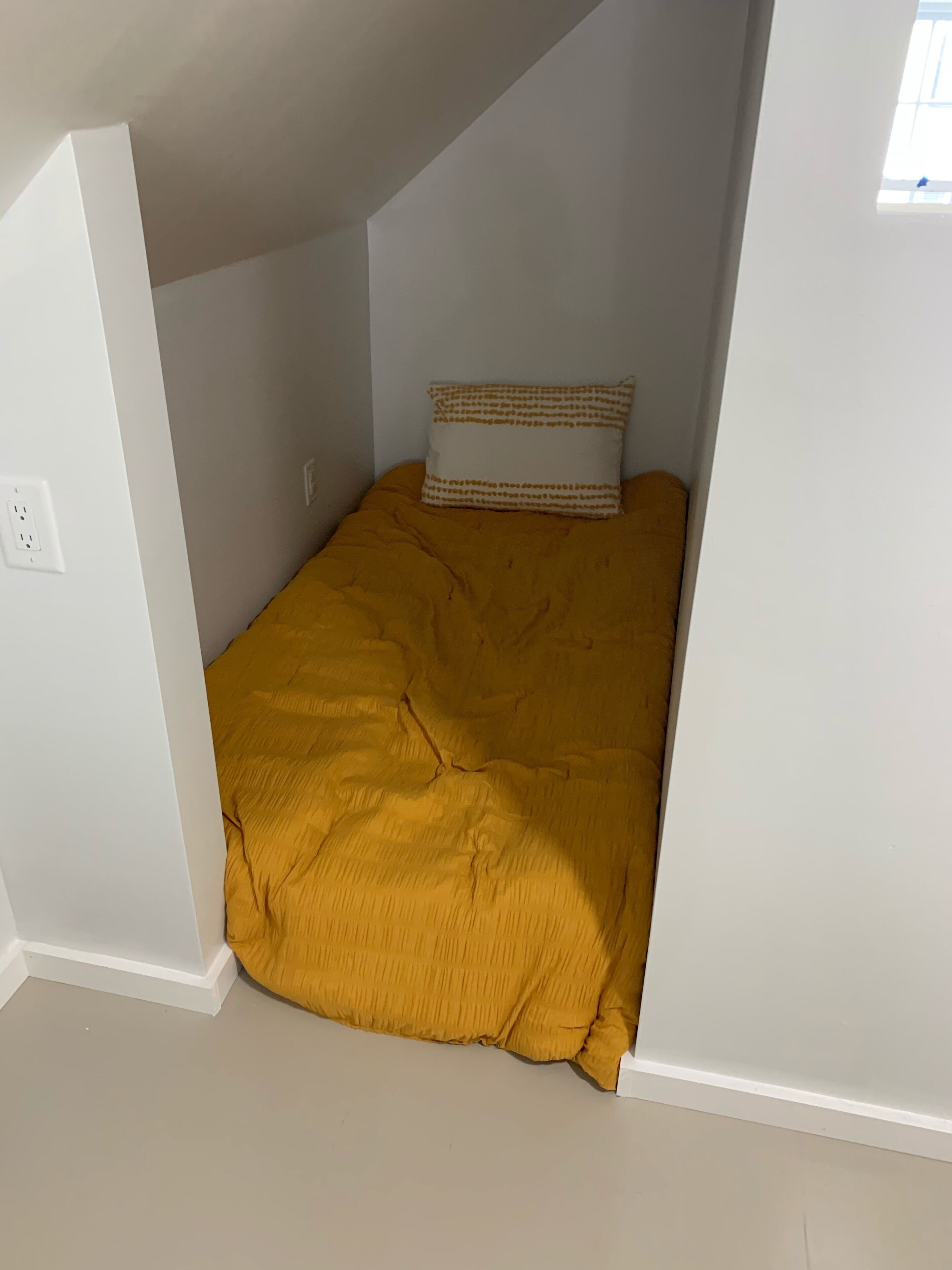 Ein kleines Zimmer mit einem Einzelbett, einer gelben Tagesdecke und einem gemusterten Kissen.