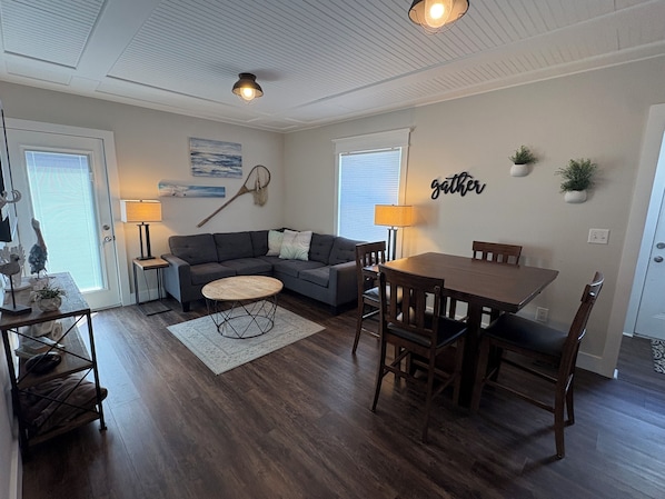 A living room with a sofa, a round coffee table, a dining table with chairs, a lamp, and a wall-mounted word 'gather'.