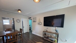 A dining area with a wooden table and chairs, a television mounted on the wall, and a hallway leading to another room.