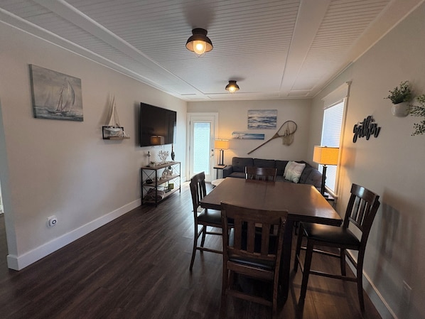 A dining area with a wooden table and chairs, a television mounted on the wall, and a ceiling light fixture.