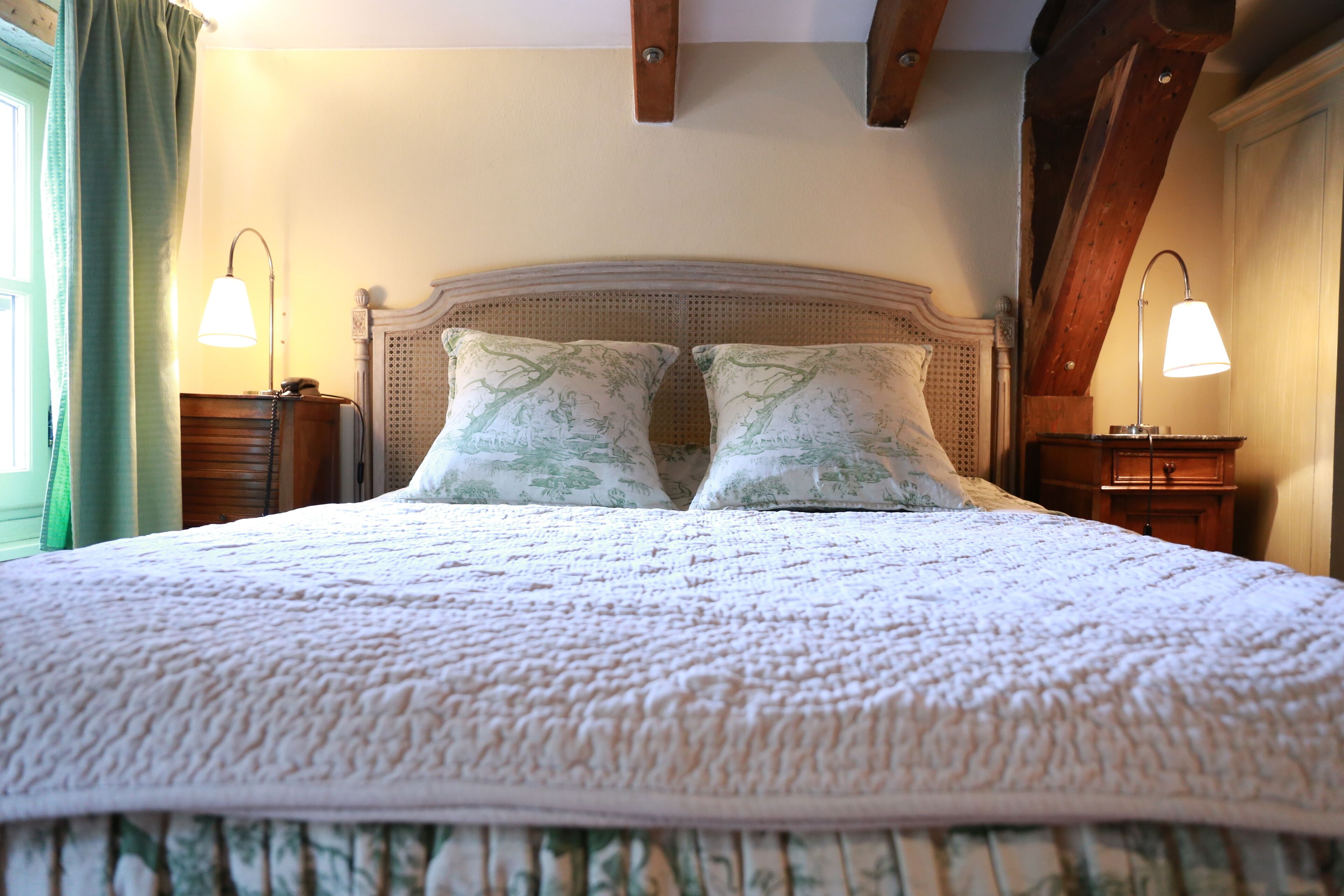 A neatly made bed with a patterned bedspread and two matching pillows, flanked by bedside tables with lamps, in a room with exposed wooden beams and a window with curtains.
