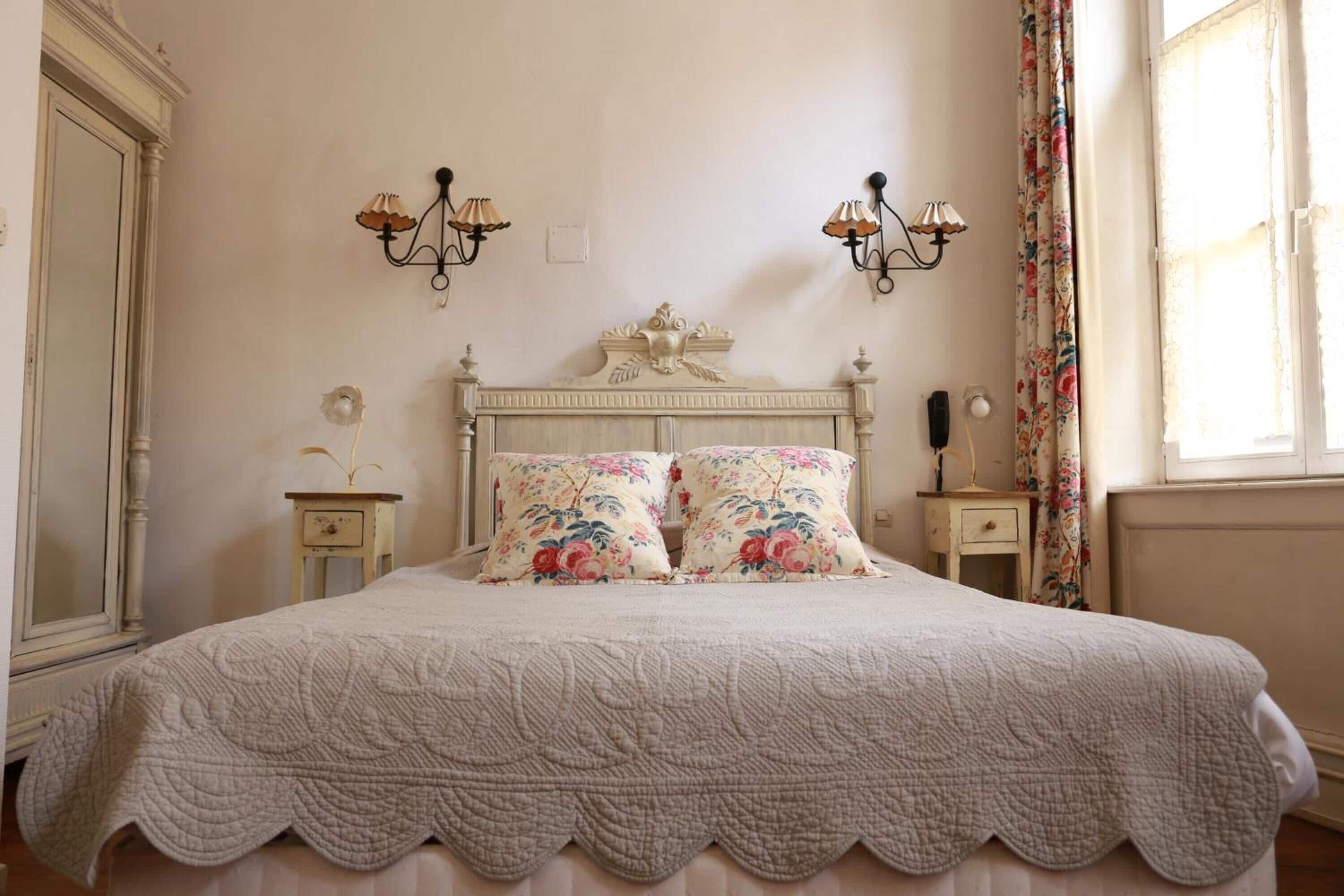 A bedroom with a large bed, floral bedding, two wall sconces, and a classic dresser.