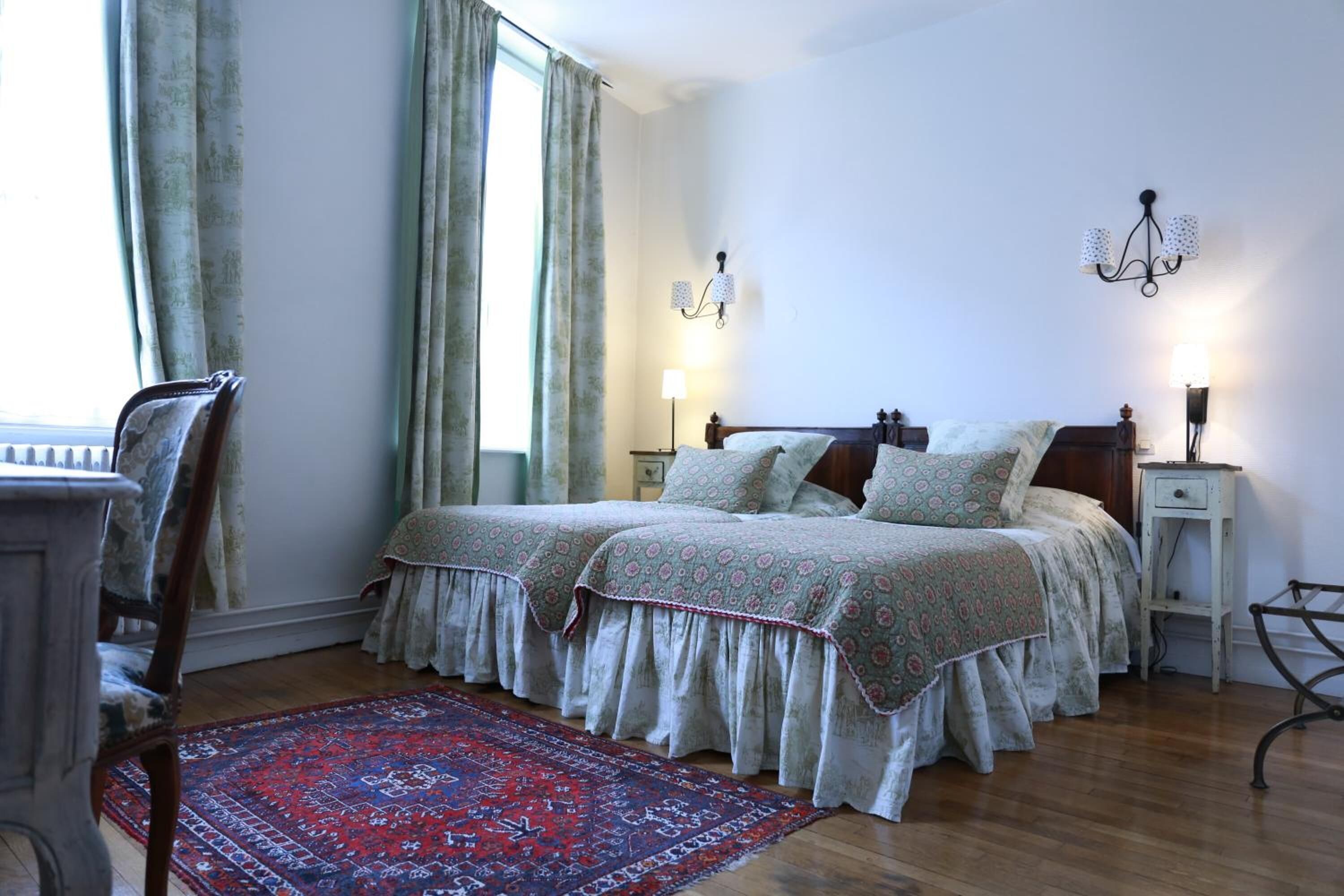 A bedroom with two beds, a wooden floor, a patterned rug, and a desk with a chair.