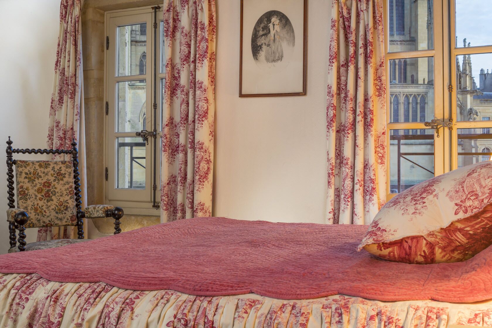 A bedroom with a bed, floral curtains, a chair, and a window with a view of buildings.