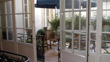 A sunlit patio with a table and chairs, a blue umbrella, and a view of the garden through a glass door.