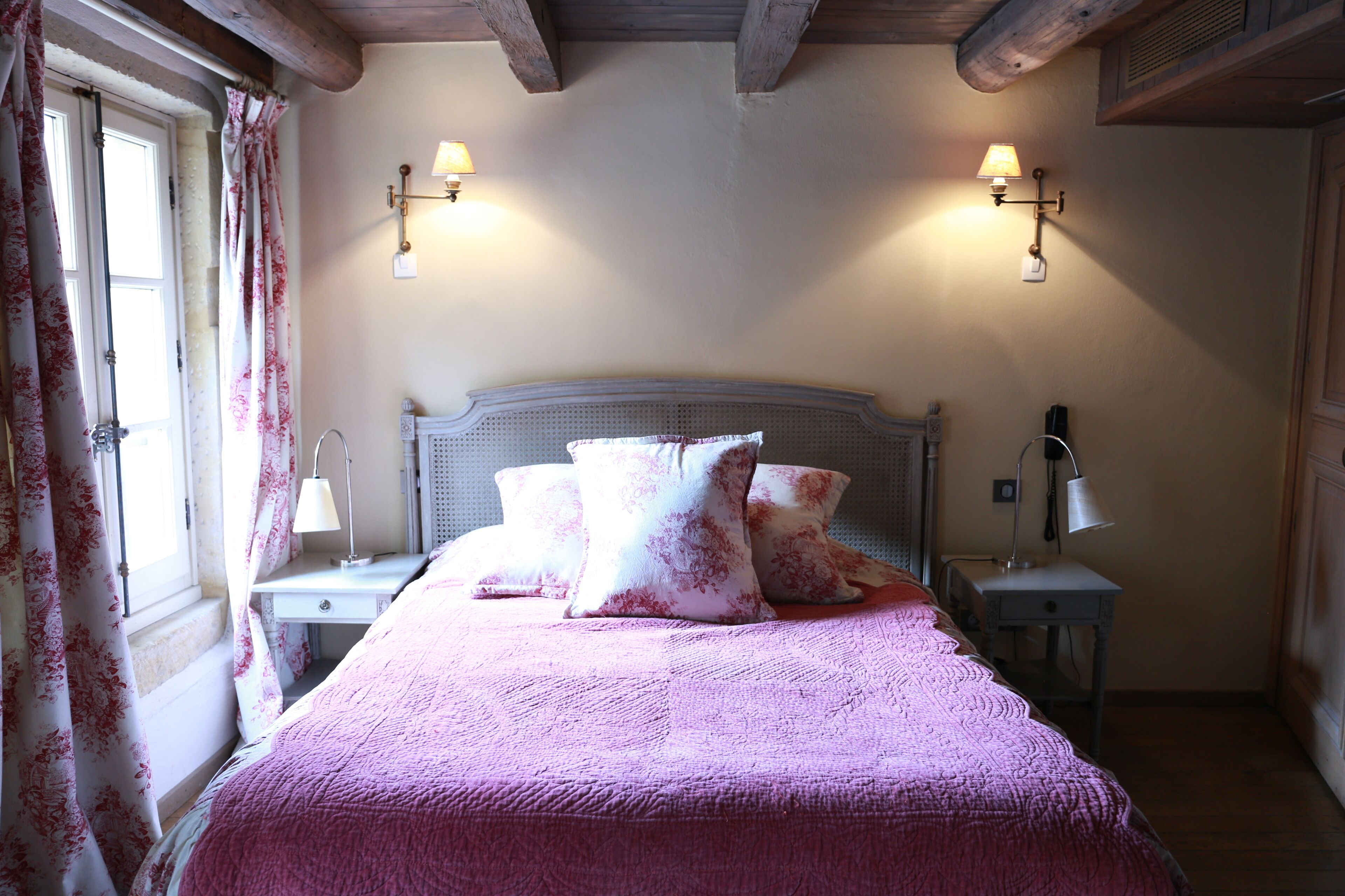 A bedroom with a bed, two bedside tables, a nightstand, a window with curtains, and two wall-mounted lamps.