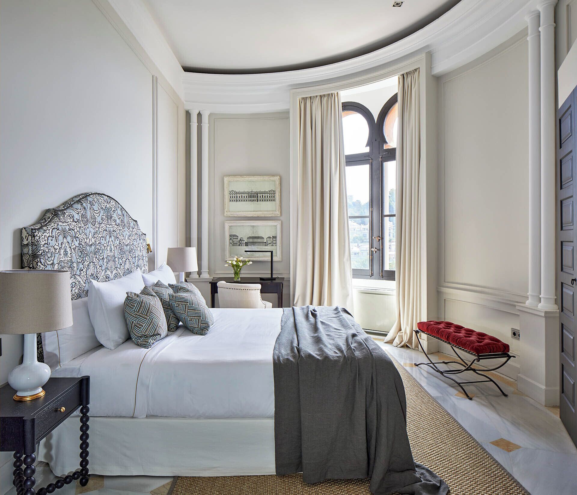 Un dormitorio con una cama grande, una mesita de noche, un banco rojo y una ventana con cortinas.