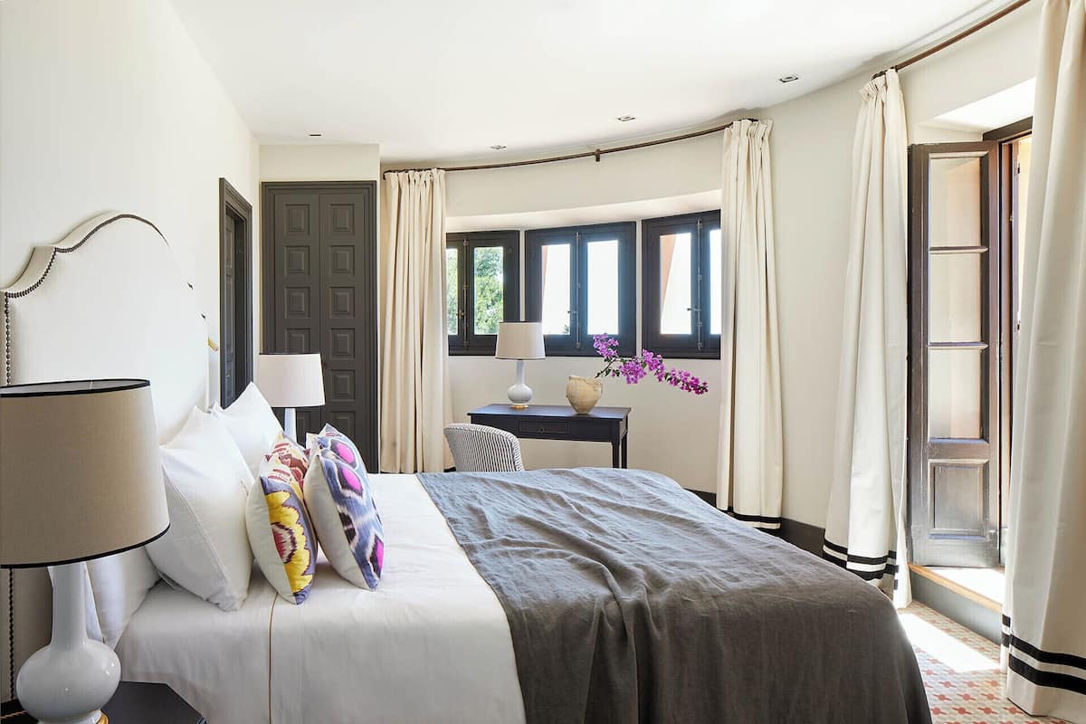 A neatly arranged bedroom with a bed, bedside table, lamp, and a view of the outdoors through windows.