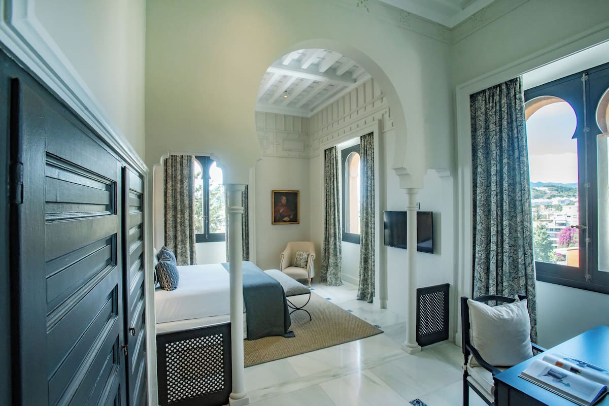 A bedroom with a bed, a chair, a desk, and a view of the city through large windows.