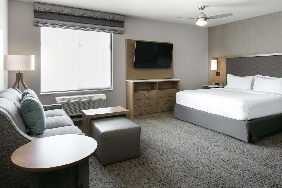 A modern hotel room with a large bed, a sofa, a small table, a TV, and a ceiling fan.