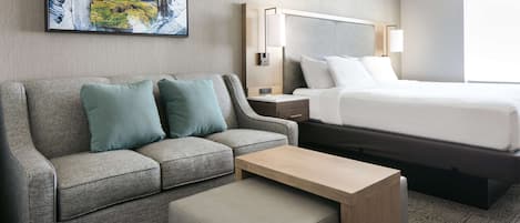 Een hotelkamer met een grijze bank, een houten bijzettafeltje, een bed met witte lakens, een nachtkastje met een lamp en een groot schilderij aan de muur.
