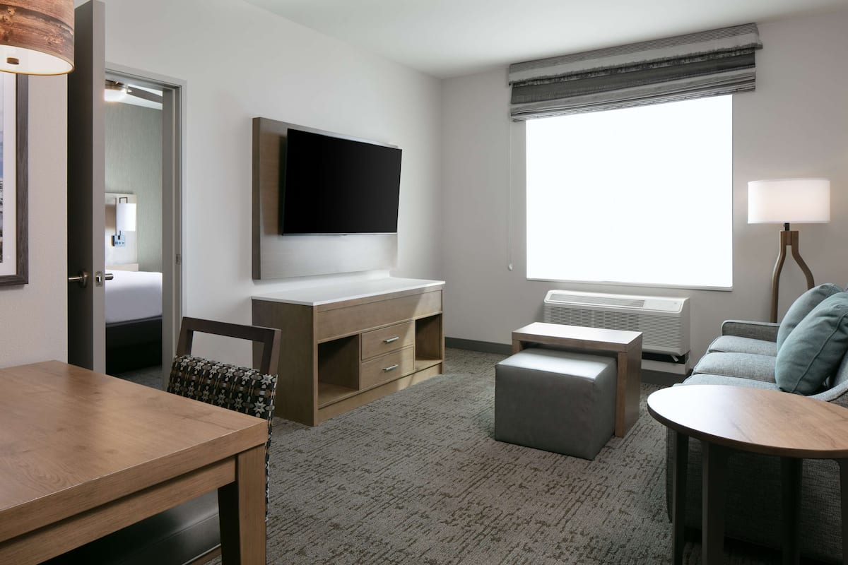 A hotel room with a wooden desk, a television mounted on the wall, a grey sofa, and a window with a view.