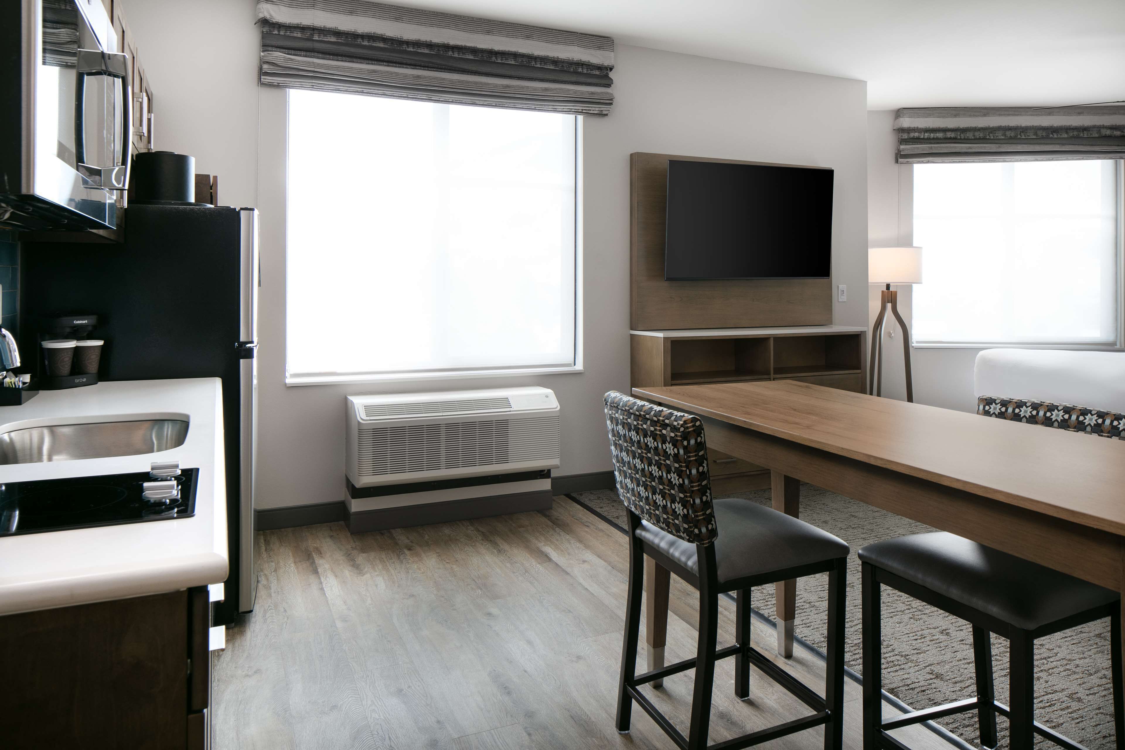 Ett modernt hotellrum med ett litet kök, ett matbord med två stolar, en platt-TV som är monterad på väggen och ett stort fönster som släpper in naturligt ljus.