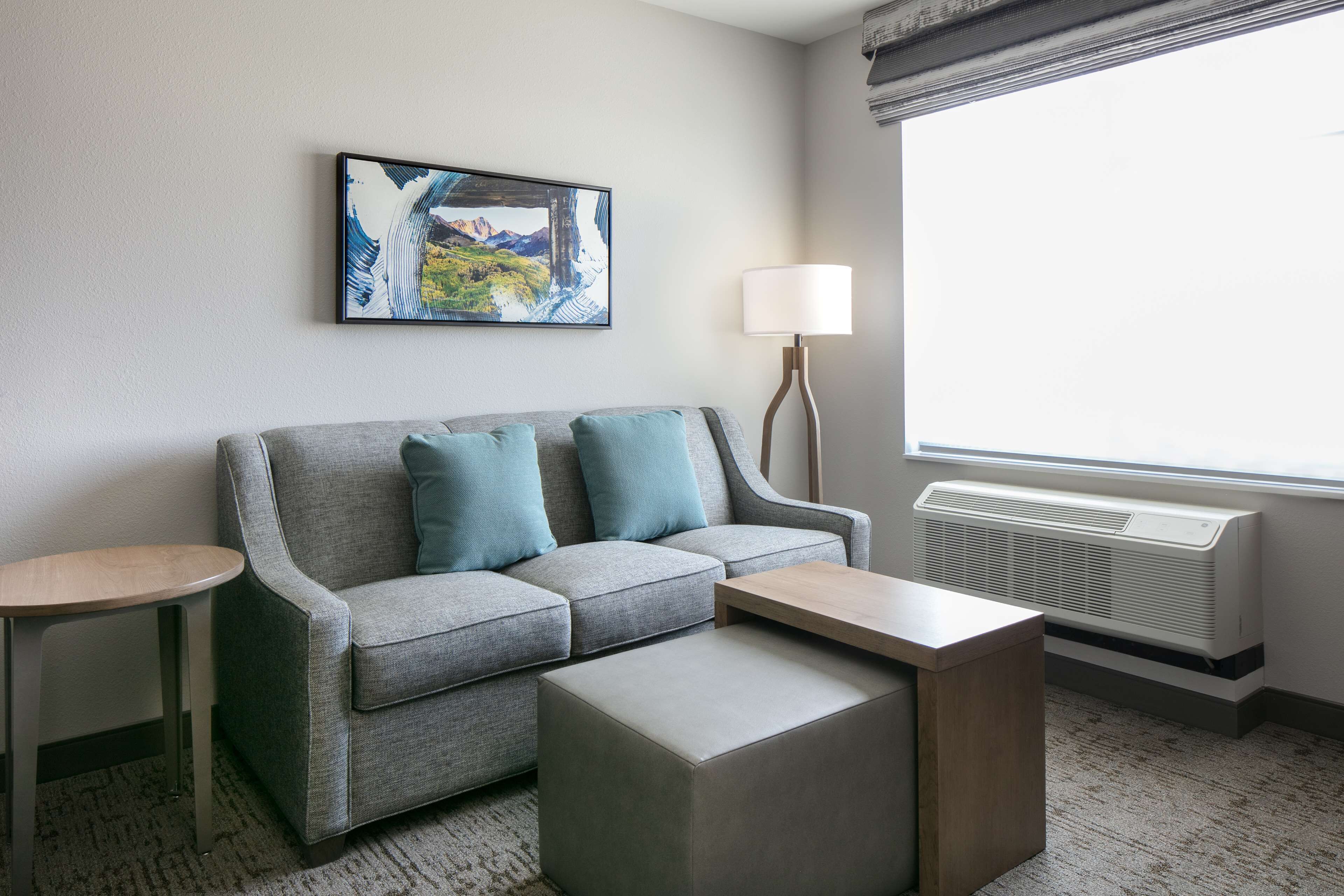Ein modernes Hotelzimmer mit grauem Sofa, zwei passenden Kissen, einem Holztisch, einem Beistelltisch, einer Stehlampe und einem großen Fenster mit Bergblick.