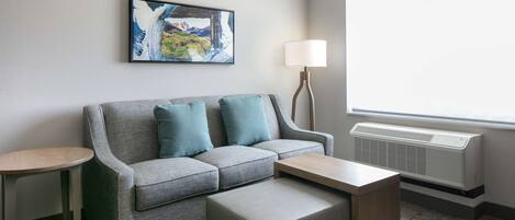 A modern hotel room with a grey sofa, two matching cushions, a wooden coffee table, a side table, a floor lamp, and a large window with a view of mountains.