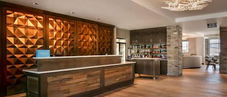 Un moderno vestíbulo de hotel con un mostrador de recepción de madera, un minibar, un pilar de piedra y una lámpara de araña.