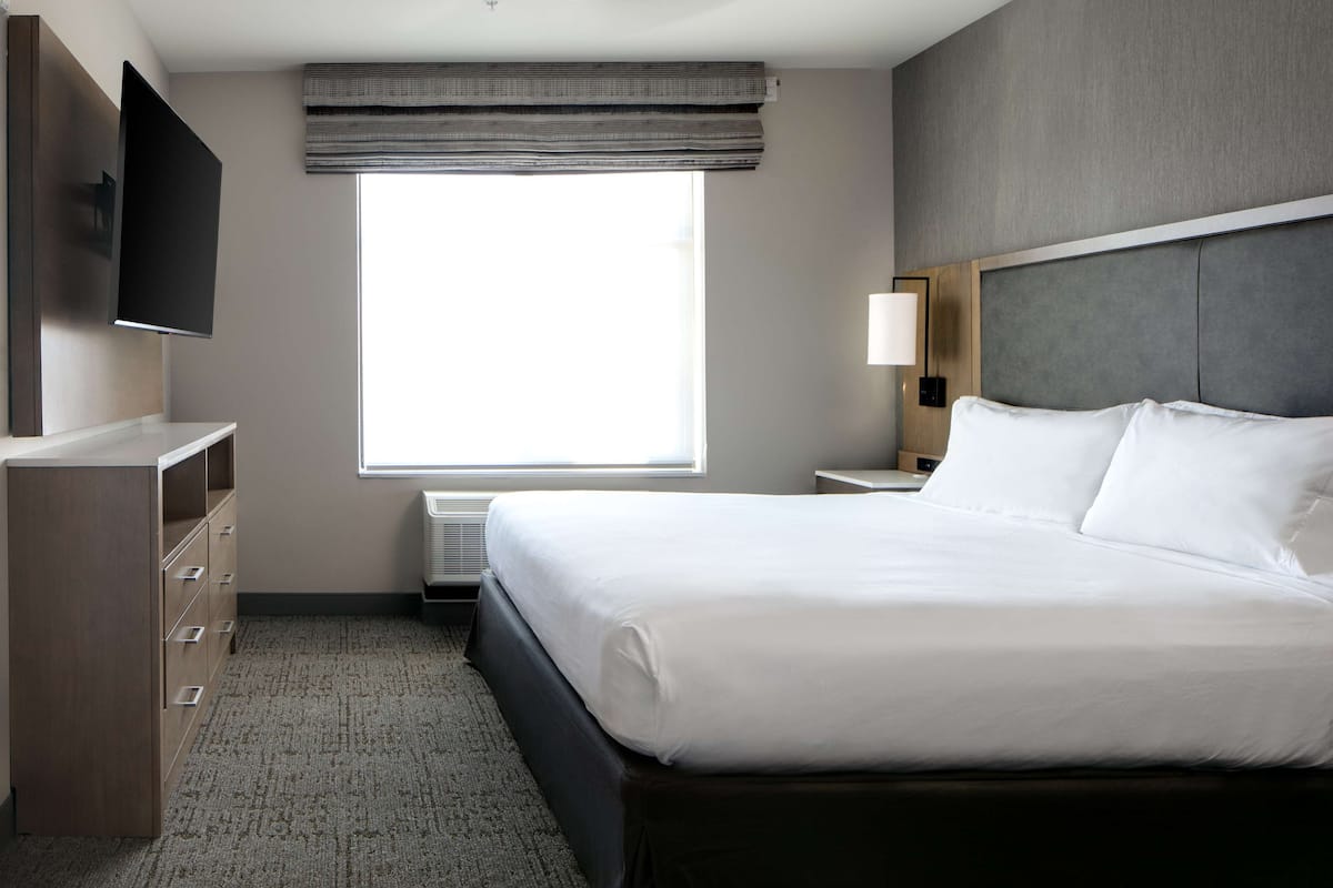 A hotel room with a large bed, a flat-screen TV mounted on the wall, a bedside table with a lamp, and a window with a view.