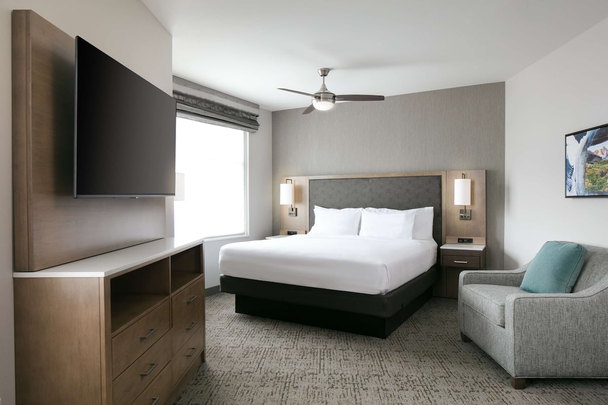 A modern hotel room with a large bed, a flat-screen TV mounted on the wall, a ceiling fan, and a view of the mountains through the window.