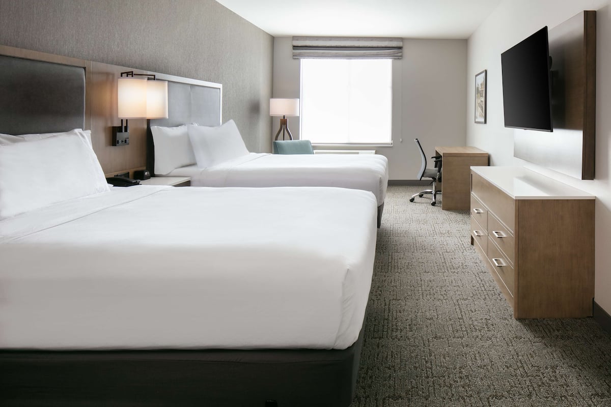 A hotel room with two beds, a desk, a chair, a TV, and a window with blinds.