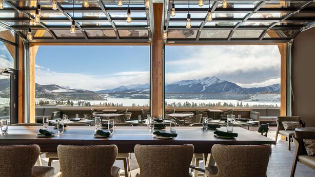 A dining area with a table set for six, large windows offering a mountain view, and a modern ceiling design with exposed beams and hanging lights. - Homewood Suites by Hilton Dillon (Dillon)