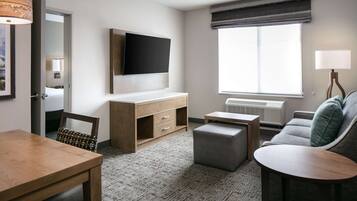 A modern hotel room with a wooden desk, a grey sofa, a flat-screen TV mounted on the wall, a large window with a view, and a small table.