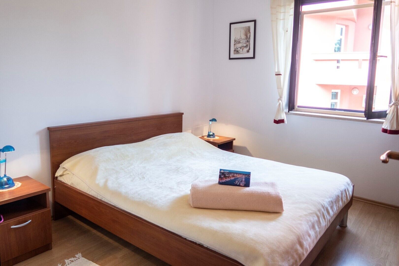 Ein ordentlich bezogenes Bett mit weißer Daunendecke und gefaltetem Handtuch, eine Nachttischlampe, ein kleiner Nachttisch mit Lampe und ein gerahmtes Bild an der Wand.