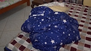 A bedroom with a bed covered by a blue blanket with star patterns, a wooden headboard, a nightstand with a bottle, and a window with curtains. - Compassionate Homestead (Kampala)