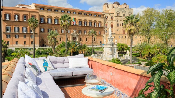 A rooftop terrace with a sofa, a table, and a view of a historic building with palm trees. - Palermo Magic House (palermo)