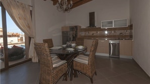 A dining area with a round table, wicker chairs, and a chandelier. A kitchen with modern appliances is visible in the background. - Casa Rural La Morisca (Tuineje)
