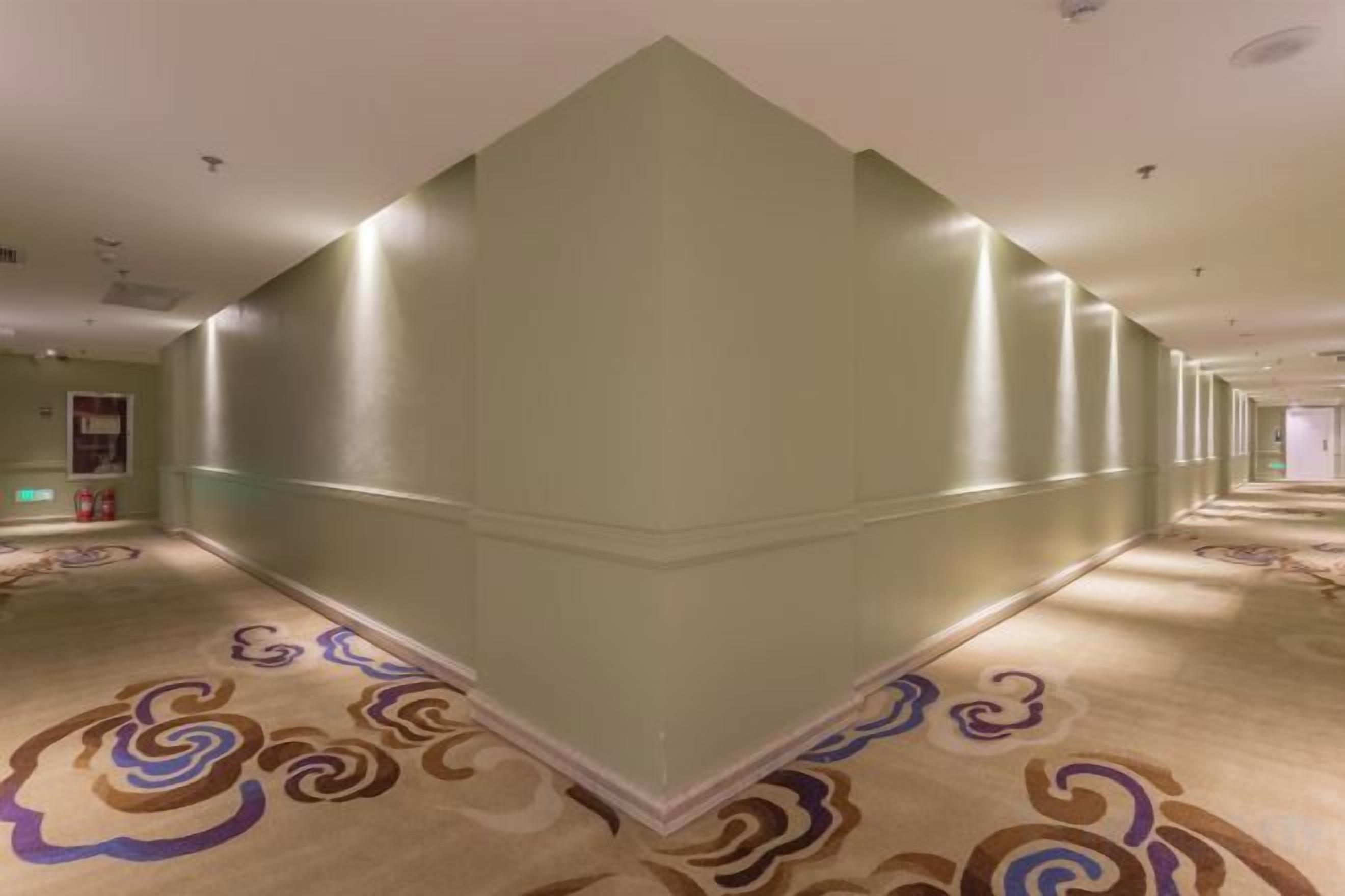 A hotel corridor with a patterned carpet, a mirrored wall, and a ceiling with recessed lighting.