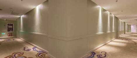 A hotel corridor with a patterned carpet, a mirrored wall, and a ceiling with recessed lighting.