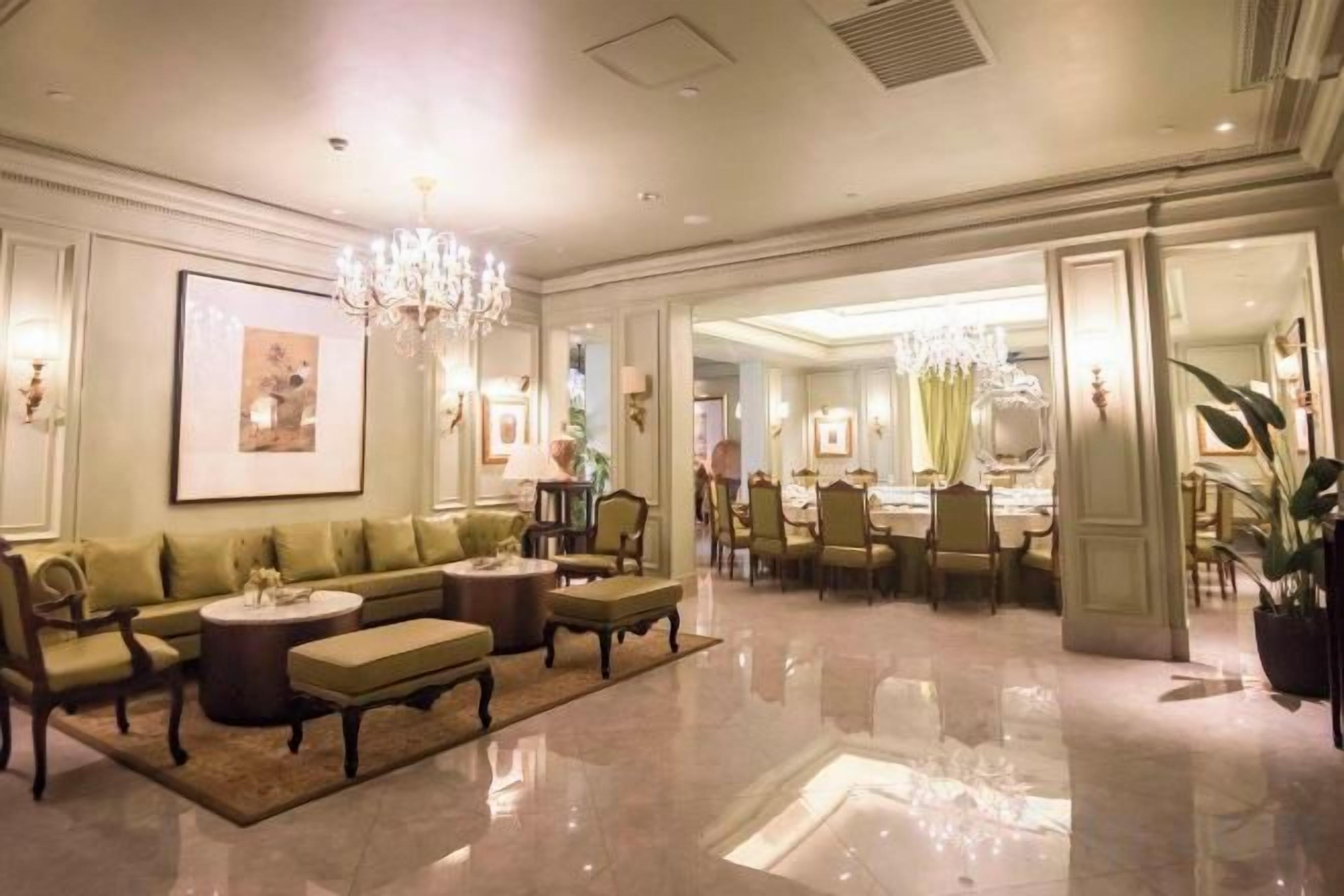 A hotel lobby with a green sofa set, a chandelier, and a dining area in the background.