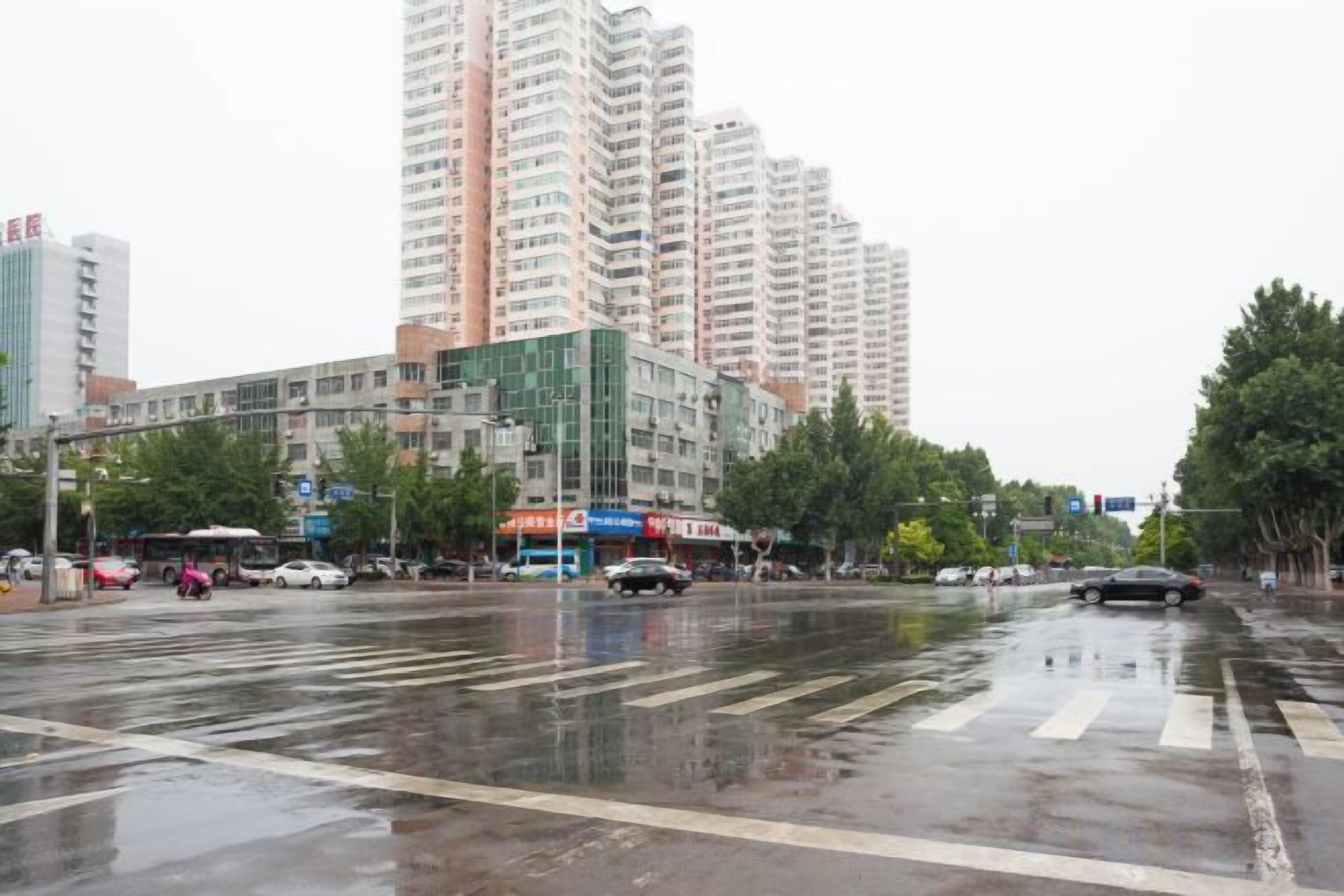 A wet city street with buildings, a crosswalk, and vehicles.
