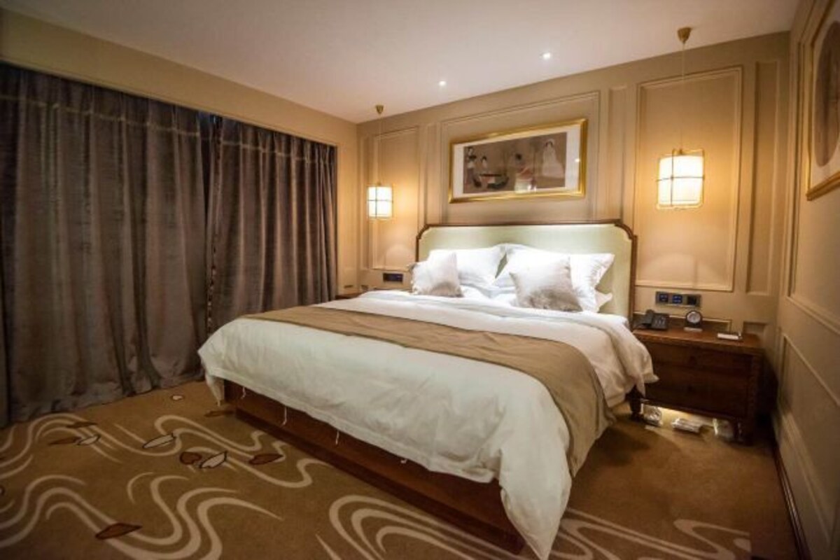 Una camera d'albergo con un letto grande, due comodini, un tavolino e una lampada a parete, con un quadro sopra il letto.