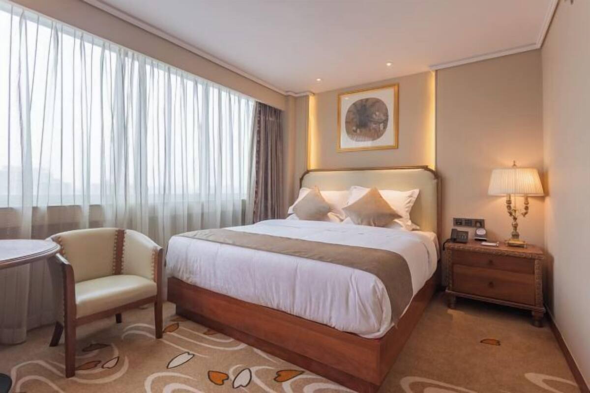 Een hotelkamer met een bed, nachtkastje, lamp, stoel en uitzicht op de stad door dunne gordijnen.
