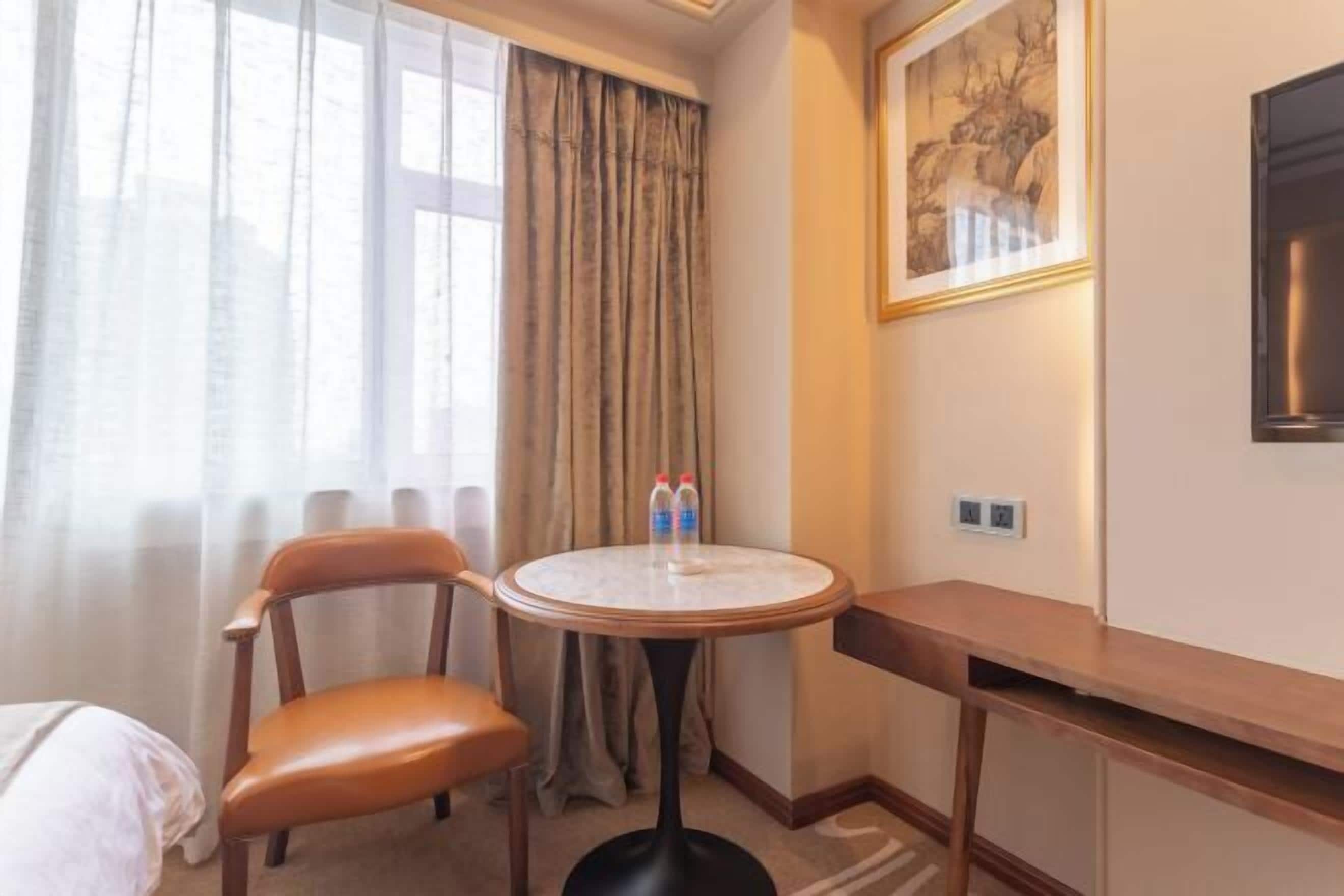 Habitación de hotel con cama, una silla, una mesa redonda con dos botellas, un cuadro y un televisor en la pared.