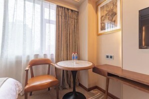 A hotel room with a bed, a chair, a round table with two bottles, a painting, and a TV on the wall. - Century Plaza Qidu Hotel (Zibo)