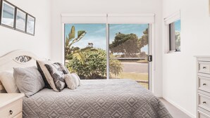 Una camera da letto con un letto, cuscini, un comodino, una cassettiera e una porta scorrevole in vetro che si apre su una vista di vegetazione e una strada.