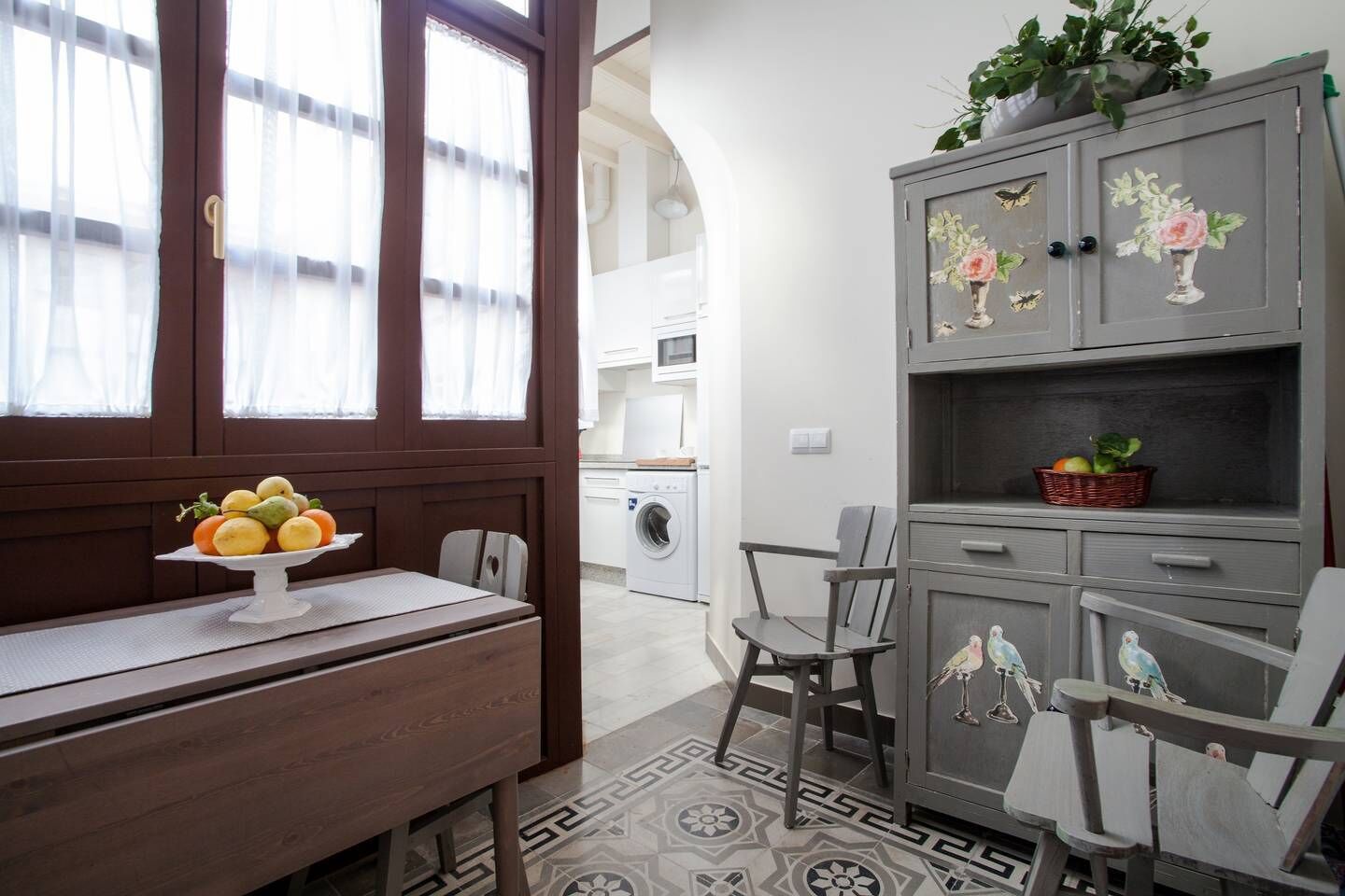 Een keuken met een houten tafel, stoelen, een presentatie van fruit en een grijs kastje met bloemversiering.