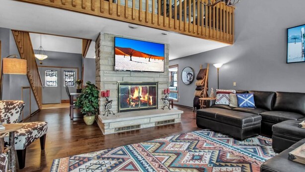 A spacious living room with a fireplace, a large TV mounted above it, and a cozy seating area with a black leather couch and a patterned rug.