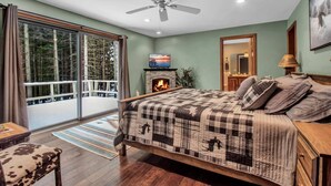 A bedroom with a large bed, a fireplace, a television, and a balcony with a view of trees.